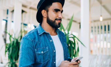 Moda blogunda yayınlanması için elinde cep telefonu cihazıyla gezen modaya uygun giyinmiş İspanyol hippinin yan görüntüsü. Elinde dijital teknoloji olan şık şapkalı olumlu bir adam.