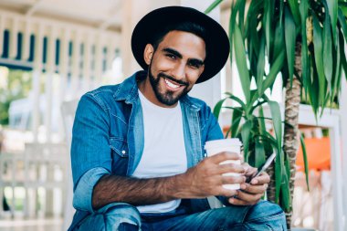 Havalı kıyafetler giymiş mutlu bir adamın portresi. Elinde yemek kupası ve akıllı telefon aleti ile dışarıda oturuyor. Boş zamanlarında kameraya gülümsüyor.