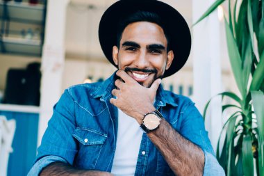 Moda kıyafetleri içinde mutlu gülümseyen bir adamın portresi ve şık siyah şapkası gençlik sokak kafeteryasının yanında dinleniyor, neşeli bir Türk genci kentte boş vakitlerin tadını çıkarıyor.