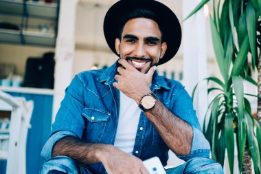 Moda kıyafetleri içinde mutlu gülümseyen bir adamın portresi ve şık siyah şapkası gençlik sokak kafeteryasının yanında dinleniyor, neşeli bir Türk genci kentte boş vakitlerin tadını çıkarıyor.