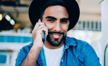 Siyah şapkalı, kameraya gülümseyen ve arkadaşlarıyla cep telefonu iletişiminin keyfini çıkaran pozitif adamın portresini kapat. Operatöre telefon etmek için kablosuz bağlantıyı kullanan neşeli bir genç.