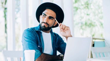 Düşünceli Türk değişim öğrencisi, üniversite kafeteryasında modern dijital dizüstü bilgisayar ve uzaktan çalışma konseptiyle sınav savunması için dil öğrenme üzerine kafa yoruyor.