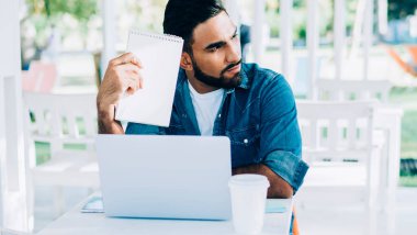 Tasarım derslerinden ders çizimleri alan ve online proje için yeni fikirler düşünen düşünceli bir Türk öğrenci, açık havada öğrenim gören düşünceli bir adam.