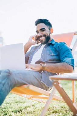 Mutlu hipster adam cep telefonu uygulaması ile dostça uluslararası sohbetten zevk alıyor dışarıda oturuyor ve bilgisayarla uzaktan çalışıyor, neşeli adam çağrı sırasında seyahat planlarını tartışıyor ve ceza kesiyor.