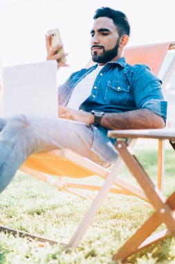 İspanyol erkek serbest çalışan akıllı telefon ve dizüstü bilgisayarla medya dosyalarını paylaşıyor bluetooth bağlantısını açık havada kullanıyor, yetenekli hippi adam cep telefonu aygıtıyla e-posta mesajlarını kontrol ediyor.