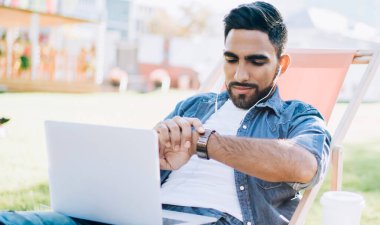 Dizüstü bilgisayar teknolojisiyle uzaktan yakından ilgilenirken takılabilir kol saatini kontrol eden yetenekli erkek serbest yazar, arkadaşı beklerken elektronik kulaklıkla müzik dinleyen genç Türk adam