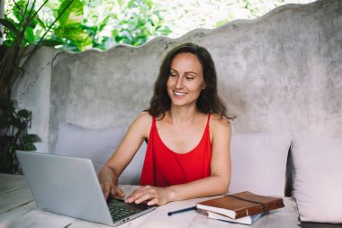 Kırmızı elbiseli, gülümseyen orta yaşlı bir kadın. Parlak, bitki dolu bir kafede dizüstü bilgisayarla çalışıyor. Freelancer rahat ve verimli bir atmosferin keyfini çıkarıyor. Yanında da defter var..