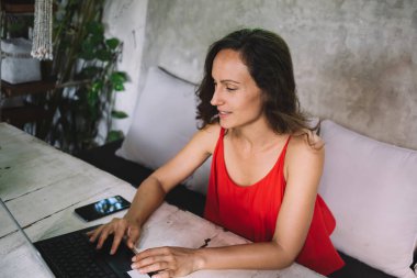 Kırmızı elbiseli, orta yaşlı bir kadın şık, bitkilerle dolu bir kafede dizüstü bilgisayarla çalışıyor. Çevresi yeşillik, akıllı telefon ve kırsal kesimle çevrili serbest çalışan bir profesyonel. Sakin,