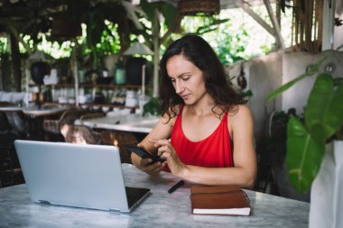 Odaklanmış orta yaşlı, kırmızı elbiseli bir kadın, verimli, bitki dolu bir kafede dizüstü bilgisayarla çalışırken akıllı telefon kullanıyor. Çevresi yeşillik, defter ve huzur ile çevrili bir serbest yazar.,
