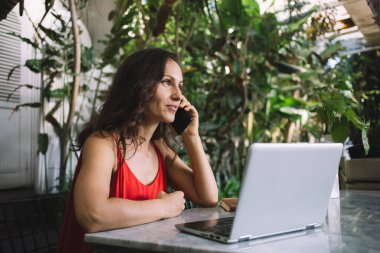 Kırmızı elbiseli, orta yaşlı bir kadın yemyeşil bir kafede dizüstü bilgisayarla uğraşırken telefonla konuşuyor. Doğadan esinlenilmiş dekorla çevrili