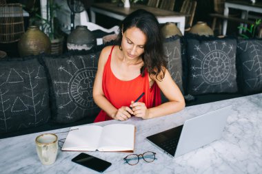 Laptop, açık defter, bardak ve kahve ile mermer bir masada çalışan kırmızı elbiseli serbest çalışan biri. Dekoratif yastıklı rahat dış mekan, üretken ve rahat bir ortam yaratıyor.