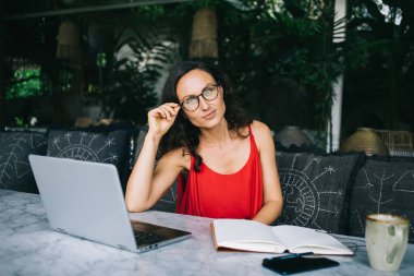 Kırmızı elbiseli serbest çalışan, dizüstü bilgisayar üzerinde çalışırken gözlük takan biri. Açık bir defter ve kahveyle mermer bir masada oturuyor, rahat ve üretken bir atmosfer yaratıyor.