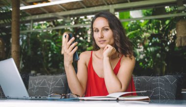 Kendine güveni tam bir kadın elinde akıllı bir telefonla tropik bir ortamda dizüstü bilgisayarla çalışıyor. Bereketli yeşilliklerle çevrili, odaklanmış ve meşgul görünüyor, sakin bir çalışma ortamının tadını çıkarıyor.