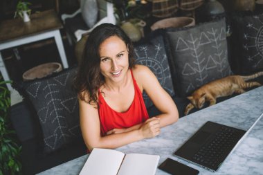 Rahat ve şık bir ortamda dizüstü bilgisayar ve defterle uzaktan çalışan gülümseyen bir kadın. Kızıl bir kedi yakınlarda dinleniyor, üretken çalışma alanına rahat bir dokunuş serpiştiriyor.