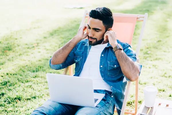 Elektronik kulaklıklar ve dizüstü bilgisayarla müzik dinleyen yakışıklı bir adam, üniversite kampüsünde öğrenim görürken ses kitabına odaklanmış genç bir adam.
