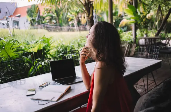 Kırmızı elbiseli serbest çalışan, kahvenin tadını çıkaran, özenle yemyeşil tropikal bahçeye bakan biri. Masadaki dizüstü bilgisayar, telefon ve defter uzaktan kumandayı gösteriyor. Barışçıl, ilham verici çalışma alanı