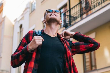 Güneşli bir günde şehir caddesinde yürürken sırt çantası ayarlarken gülümseyen ve yukarı bakan şık giysili yakışıklı bir adam.
