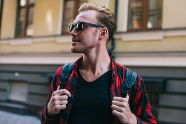 Güneş gözlüklü ve sırt çantalı yakışıklı adam güneşli bir günde şehrin ortasında yol kenarında dikilirken gözlerini kaçırıyor.