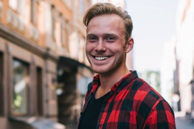 Kareli gömlekli yakışıklı adam neşeli bir şekilde gülümsüyor ve güneşli bir günde şehir caddesinde dikilip kameraya bakıyor ve modern akıllı telefonlara bakıyor.