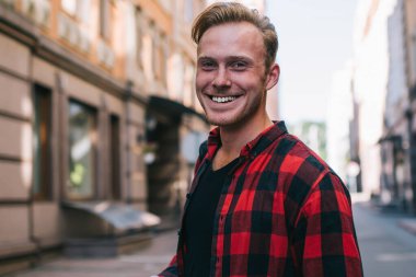 Kareli gömlekli yakışıklı adam neşeli bir şekilde gülümsüyor ve güneşli bir günde şehir caddesinde dikilip kameraya bakıyor ve modern akıllı telefonlara bakıyor.