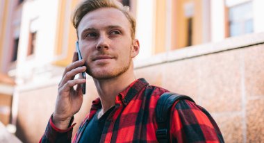 Kareli gömlekli yakışıklı adam güneşli bir günde şehir sokağının bulanık arka planında dikilirken cep telefonuyla konuşuyor ve yüzünü çeviriyor.