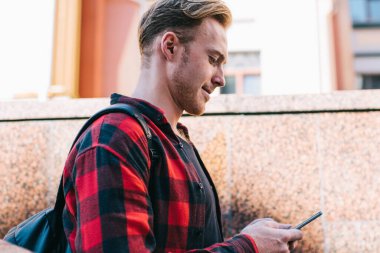 Güneşli bir günde şehir caddesinde yürürken modern akıllı telefondan mesaj yazan kareli tişörtlü yakışıklının yan görüntüsü