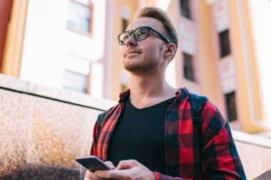 Şık gözlükler takmış yakışıklı bir adam akıllı telefondan mesaj yazarken ve güneşli bir günde şehir sokağında dikilirken başka tarafa bakıyor ve düşünüyor.