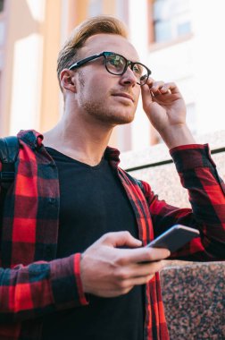 Modern akıllı telefonlu yakışıklı adam şık gözlükleri ayarlıyor ve güneşli bir günde şehir caddesinde yürürken mesafeye bakıyor.