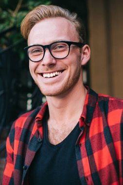 Yakışıklı genç adam günlük kıyafetlerle ve gözlüklerle neşeli bir şekilde gülümsüyor ve kameraya bakıyor.