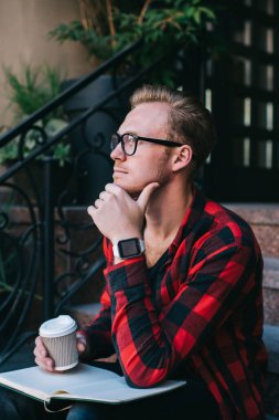 Elinde bir fincan sıcak içecekle yakışıklı bir adam ve açık bir not defteri. Çenesini ovuşturup, evin önünde oturmuş düşünürken başka tarafa bakıyor.