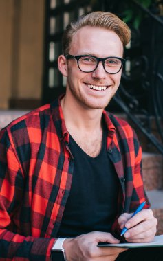 Gözlüklü ve kareli gömlekli şık bir erkek. Neşeli bir şekilde gülümsüyor ve kameraya bakıyor. Evin yakınındaki basamaklarda oturuyor ve not defteriyle yazıyor.