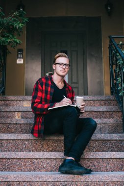 Yakışıklı bir adam şık giyinmiş ve elinde bardakla elinde sıcak bir bardak içki tutarken ve not defterine not düşerken evin önünde oturmuş kameraya bakarken.