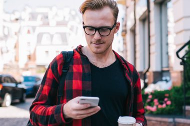 Gözlüklü şık bir adam elinde bir bardak sıcak içecek ve güneşli bir günde şehir caddesinde yürürken modern akıllı telefonlara göz atıyor.