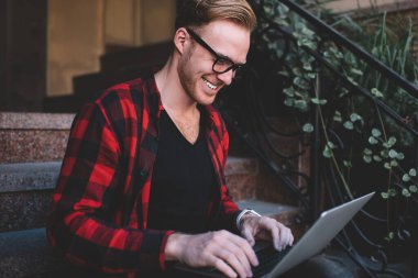 Moda gözlükleri ve damalı gömleği olan yakışıklı bir erkek. Neşeli bir şekilde gülümsüyor ve modern dizüstü bilgisayarını karıştırıyor. Şehir caddesindeki evin merdivenlerinde oturuyor.