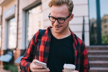 Yakışıklı adam şık kıyafetler içinde gülümsüyor ve şehir caddesinde yürürken modern akıllı telefonlara bakıyor ve sıcak içeceklerin tadını çıkarıyor.
