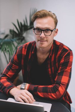 Kareli gömlekli ve gözlüklü yakışıklı. Laptop kullanıyor ve kafede otururken kameraya bakıyor.