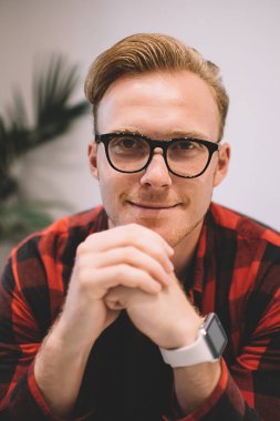 Yakışıklı, modaya uygun gözlüklü, güler yüzlü ve şık bir kafede oturmuş kameraya bakan genç adam.