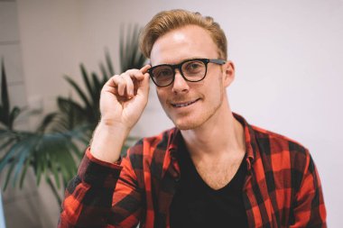 Yakışıklı, kareli tişörtlü, neşeli gözlüklü, şık bir kafede durup kameraya bakarken gülümseyen ve gözlük takan bir adam.