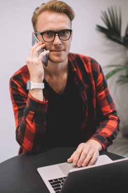 Şık gözlüklü ve kareli gömlekli zeki bir adam. Laptop kullanıyor ve kafe masasında otururken kameraya bakıyor ve akıllı telefondan konuşuyor.