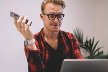 Gayri resmi giyinmiş, şık gözlükler takan, akıllı telefon tutan, kafeteryada otururken el kol hareketi yapan ve dizüstü bilgisayarla sorun yaşayan sinirli bir adam.