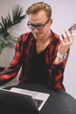 Şık giyinmiş, gözlüklü, modern akıllı telefonlu ve kafede otururken suratında asık bir ifadeyle dizüstü bilgisayara bakan yakışıklı bir adam.