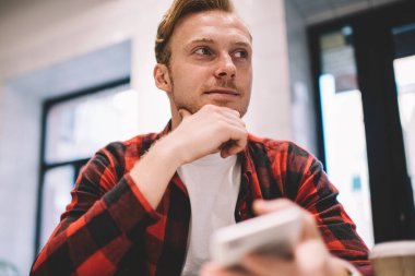 Modern akıllı telefon kullanan ve kafenin bulanık arka planında otururken yüzünü çeviren sıradan kıyafetli yakışıklı bir adam.