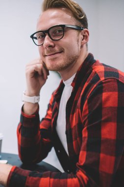 Moda gözlüklü ve kareli gömlekli yakışıklı adam şık bir kafede akıllı telefon kullanırken gülümsüyor ve kameraya bakıyor.