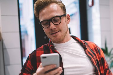 Kareli gömlekli yakışıklı bir adam elinde bir fincan sıcak içecek ve kafeteryada sandalyeye yaslanırken akıllı telefona göz atıyor.