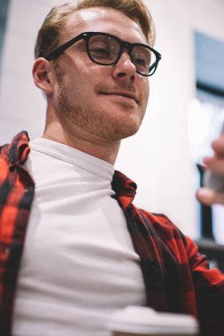 Moda gözlüklü yakışıklı ve rahat giyimli bir adam modern akıllı telefonlara bakarken şık bir kafenin bulanık arka planında rahatlıyor.