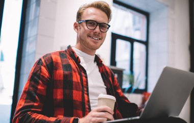 Kıyafet ve gözlüklü yakışıklı adam elinde sıcak içecek bardağı ve kafede serbest çalışmak için dizüstü bilgisayar kullanırken gülümsüyor.
