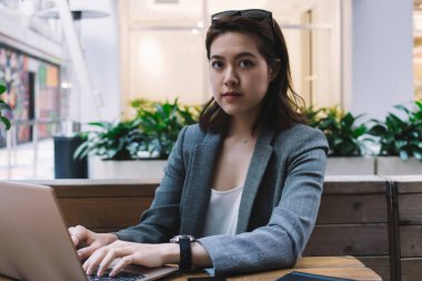 Kafede kameraya bakan akıllı telefon ve not defteriyle masada dizüstü bilgisayar kullanarak resmi giyinmiş güzel, yoğun etnik kadın serbest çalışan.