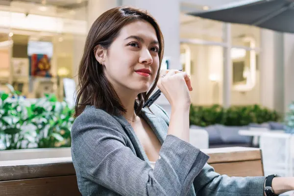 Klasik takım elbiseli, kendine güvenen ve düşünceli Asyalı bir kadın dizüstü bilgisayar ve not defteriyle ışık koridorunda oturmuş düşünceli bakışlarını başka yöne çeviriyor.