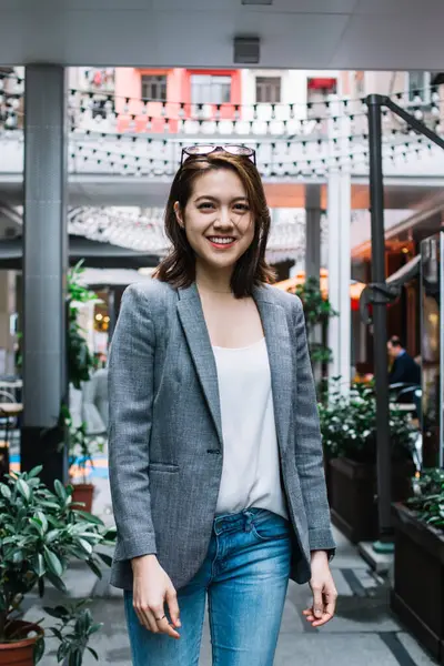 Gri ceketli, başına güneş gözlüğü takmış şark caddesinde durup kameraya gülümseyen tasasız Asyalı kadın.