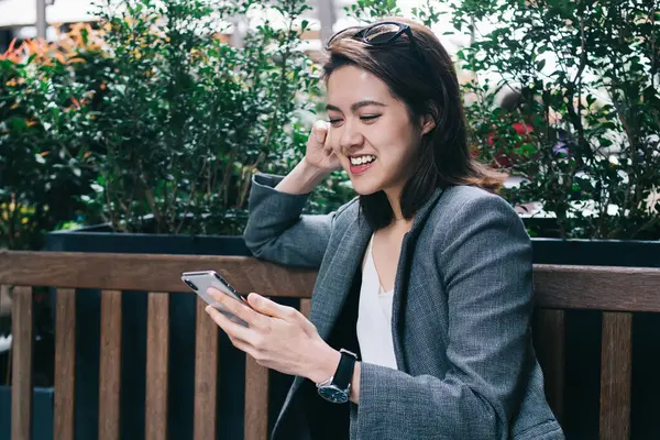 Resmi kıyafetler içinde genç, çekici, neşeli Asyalı kadın şehir sokaklarında bankta otururken modern akıllı telefonu faiziyle kullanıyor.
